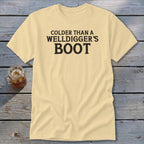 Pale yellow t-shirt with black text reading "Colder Than A Welldigger's Boot".