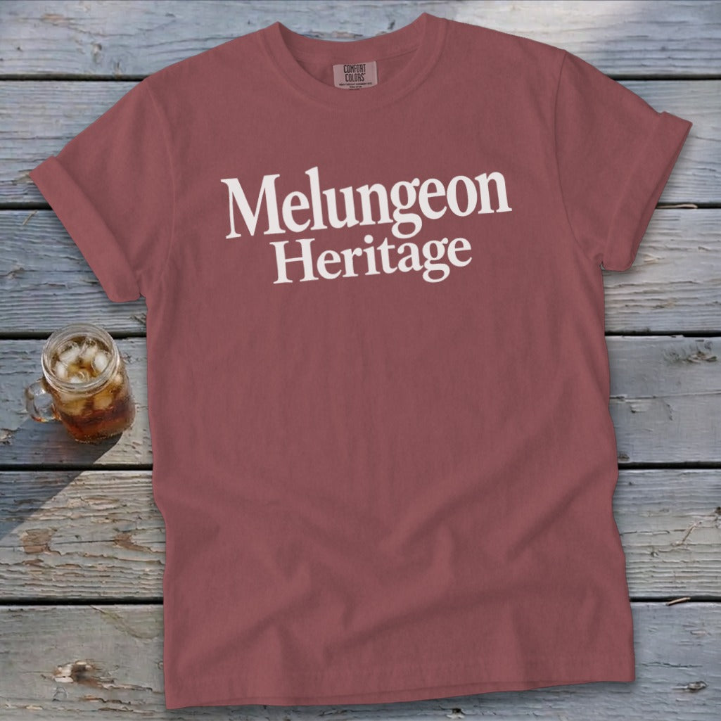 Dusty rose t-shirt with white text reading Melungeon Heritage on a weathered wooden background.