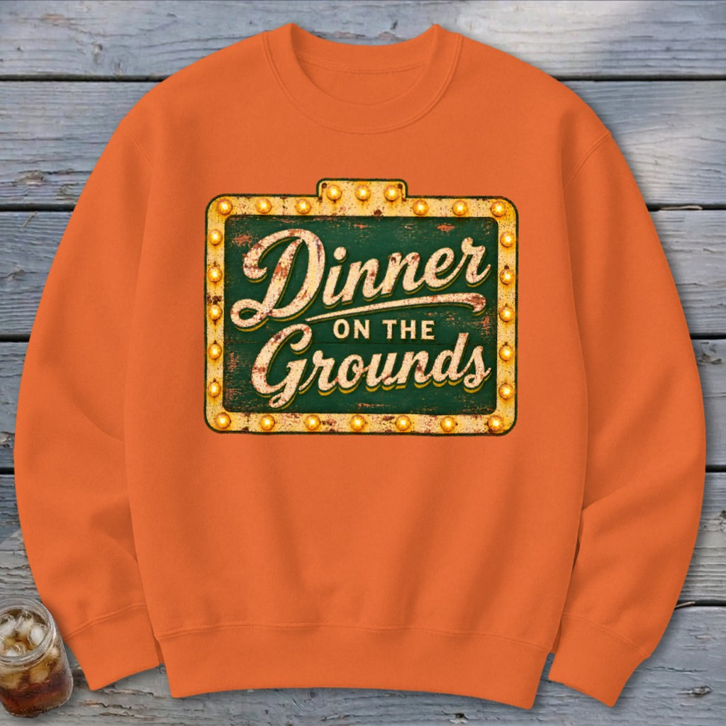 An orange crewneck sweatshirt featuring a graphic of a vintage marquee sign reading Dinner on the Grounds.