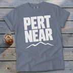A slate blue t-shirt displaying white text reading PERT NEAR above a mountain outline on a wooden surface.