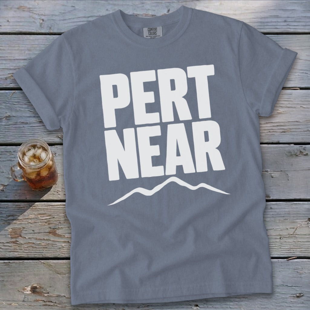 A slate blue t-shirt displaying white text reading PERT NEAR above a mountain outline on a wooden surface.