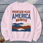 A light pink crewneck sweatshirt featuring a patriotic graphic with the text Mountain-Made America 1776-2026.