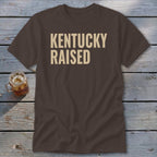 Brown t-shirt with "KENTUCKY RAISED" text in cream, next to a glass of iced tea.