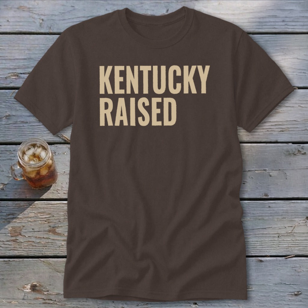 Brown t-shirt with "KENTUCKY RAISED" text in cream, next to a glass of iced tea.