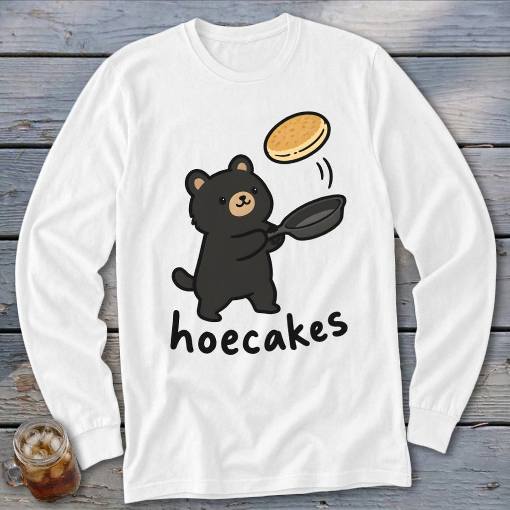 A white long-sleeved t-shirt featuring a cartoon black bear flipping a pancake in a pan, with the word hoecakes underneath.