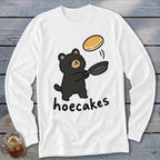 A white long-sleeved t-shirt featuring a cartoon black bear flipping a pancake in a pan, with the word hoecakes underneath.