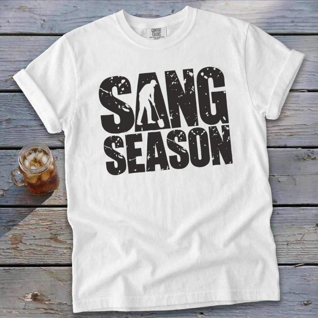 A white crewneck t-shirt with rolled sleeves lies on a rustic wooden table beside a glass of iced tea. Black distressed text on the chest reads SAND SEASON, with a silhouette of a person shoveling sand integrated into the letter A.