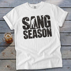 A white crewneck t-shirt with rolled sleeves lies on a rustic wooden table beside a glass of iced tea. Black distressed text on the chest reads SAND SEASON, with a silhouette of a person shoveling sand integrated into the letter A.