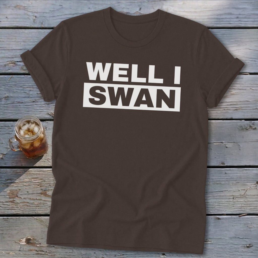 A brown crewneck t-shirt with rolled sleeves lies on a wooden surface beside a glass of iced tea, featuring the text WELL I SWAN in white uppercase letters across the chest.