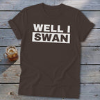 A brown crewneck t-shirt with rolled sleeves lies on a wooden surface beside a glass of iced tea, featuring the text WELL I SWAN in white uppercase letters across the chest.