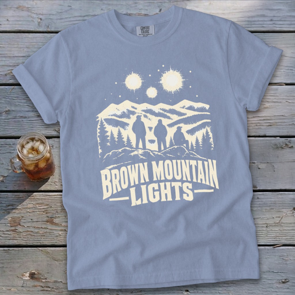 Periwinkle t-shirt featuring a white graphic of three people viewing mountain lights on a rustic wooden background.