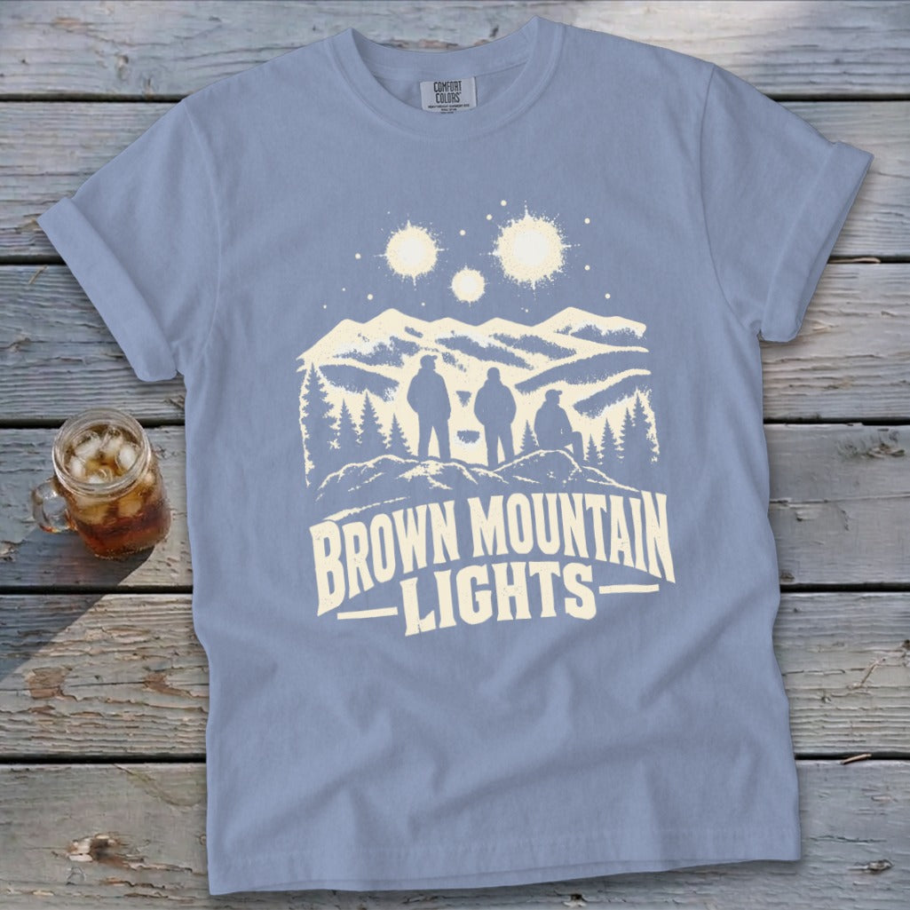 Periwinkle t-shirt featuring a white graphic of three people viewing mountain lights on a rustic wooden background.