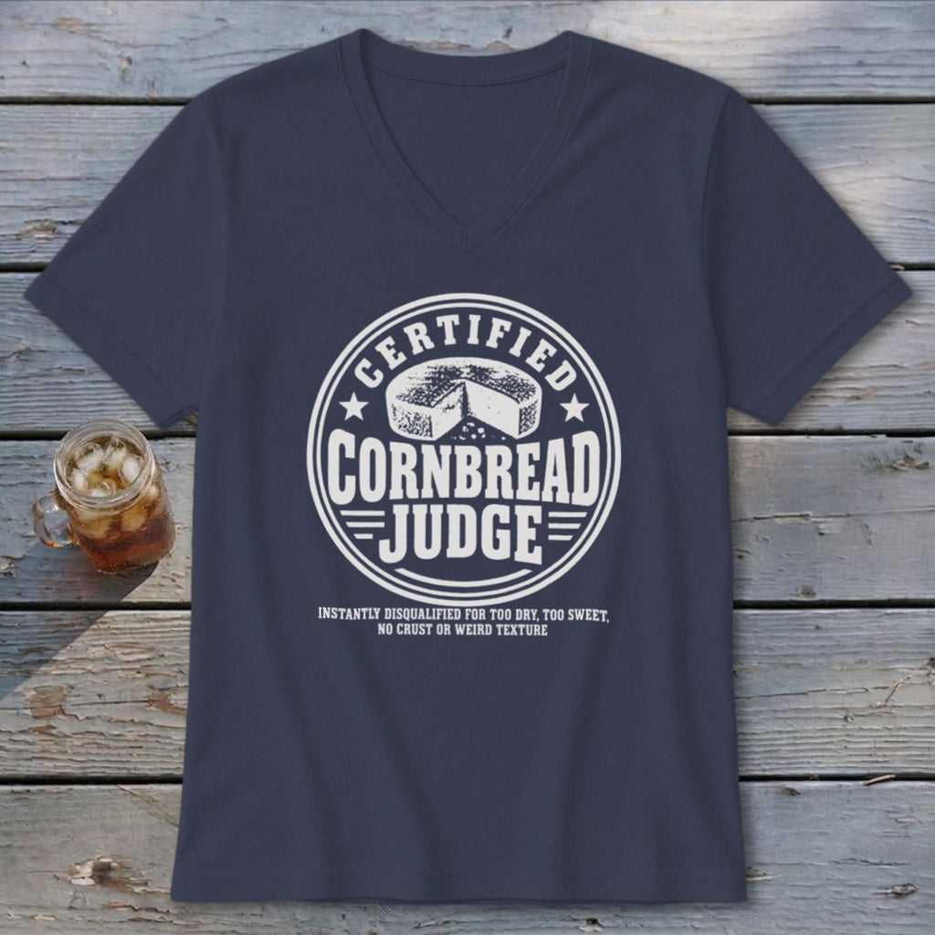 A dark blue v-neck t-shirt with a white graphic reading Certified Cornbread Judge rests on a wooden surface next to a glass of iced tea.
