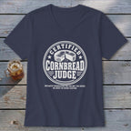 A dark blue v-neck t-shirt with a white graphic reading Certified Cornbread Judge rests on a wooden surface next to a glass of iced tea.