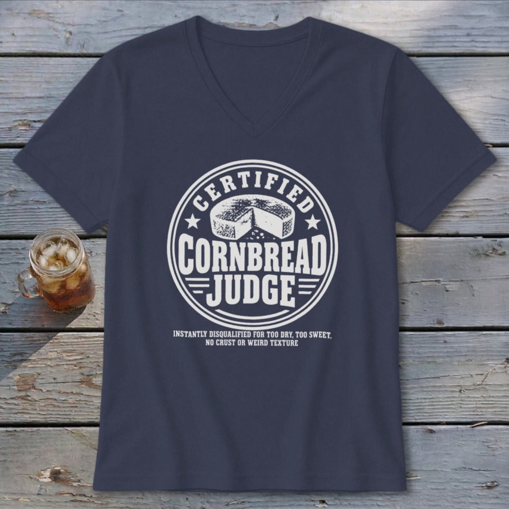 A dark blue v-neck t-shirt with a white graphic reading Certified Cornbread Judge rests on a wooden surface next to a glass of iced tea.
