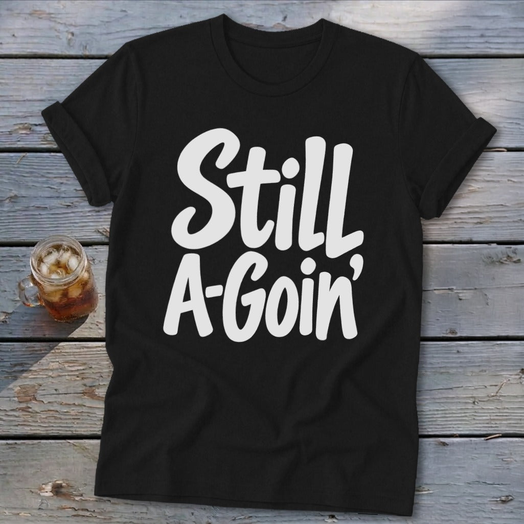 Black t-shirt featuring the white text "Still A-Goin'" laid flat on a rustic wooden surface next to an iced drink.