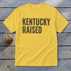 Yellow t-shirt with "KENTUCKY RAISED" text in black, beside a glass of iced tea.