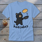 Light blue t-shirt featuring a cartoon black bear flipping a pancake with the text hoecakes.