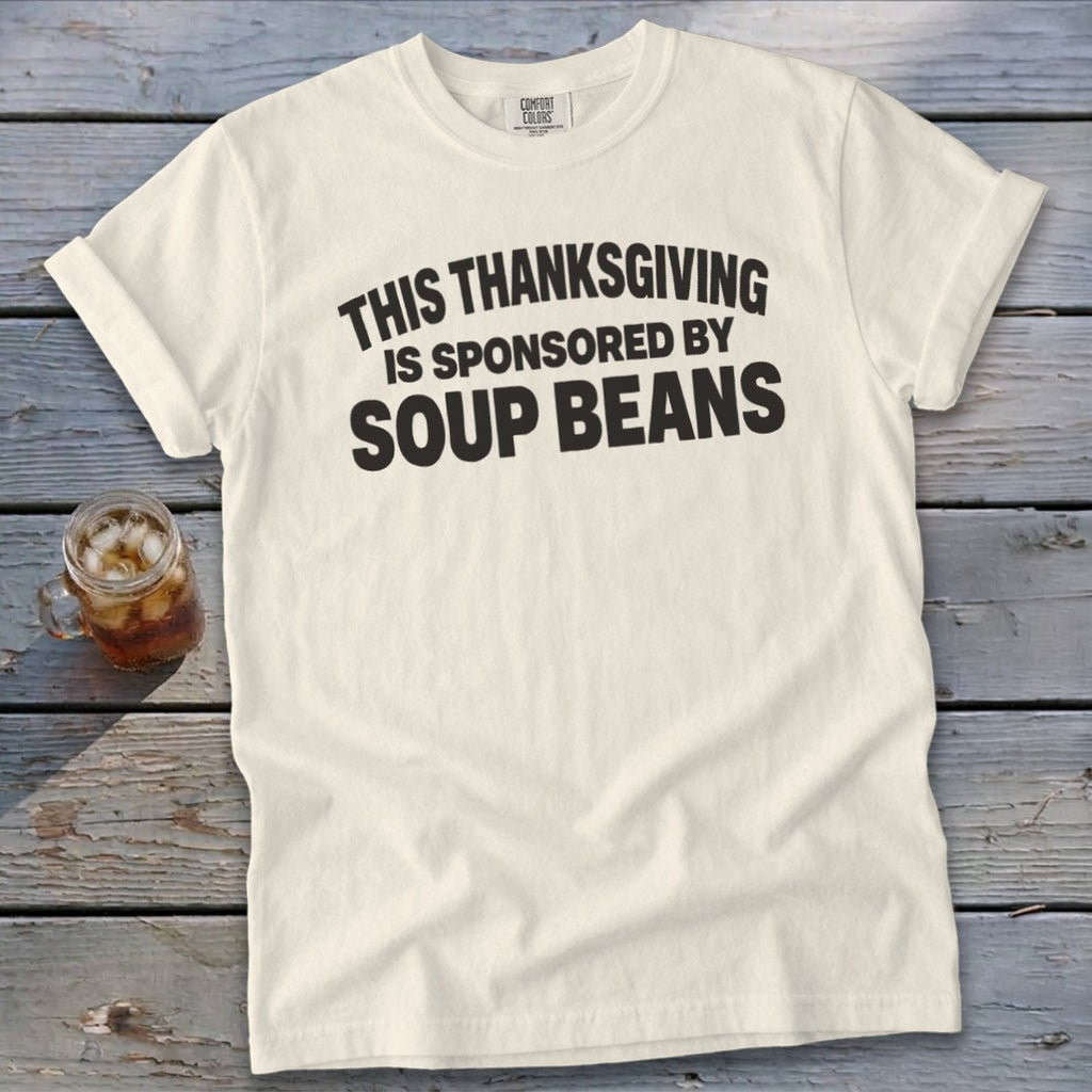 A cream-colored t-shirt with rolled sleeves lies on a rustic wooden table, featuring black text that reads THIS THANKSGIVING IS SPONSORED BY SOUP BEANS.
