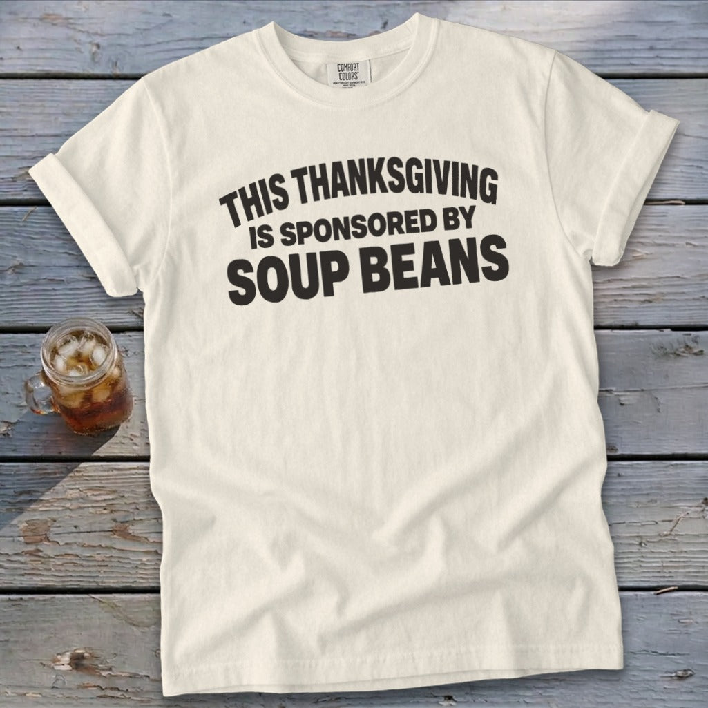 A cream-colored t-shirt with rolled sleeves lies on a rustic wooden table, featuring black text that reads THIS THANKSGIVING IS SPONSORED BY SOUP BEANS.