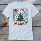 A white t-shirt features a vintage-style Christmas design with the text Mountain Merry framing a decorated tree.