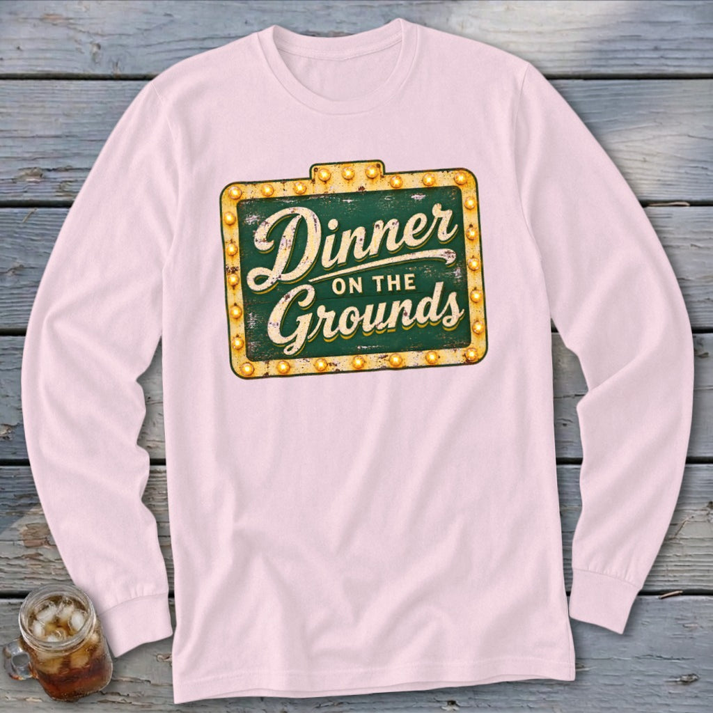 Light pink long-sleeved t-shirt featuring a green vintage sign graphic with yellow lights and text reading Dinner on the Grounds.