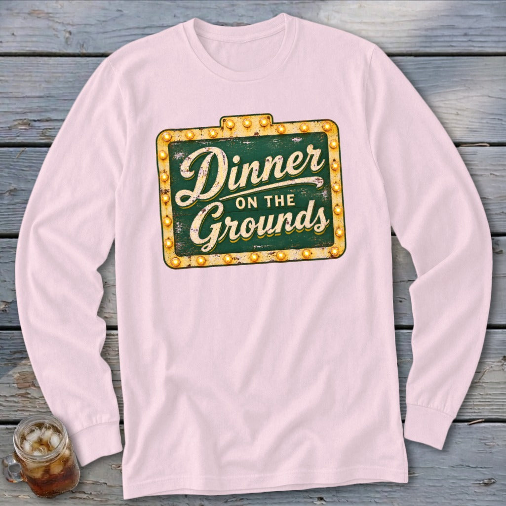 Light pink long-sleeved t-shirt featuring a green vintage sign graphic with yellow lights and text reading Dinner on the Grounds.