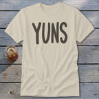 Cream t-shirt with "YUNS" in dark gray lettering on a wooden background.