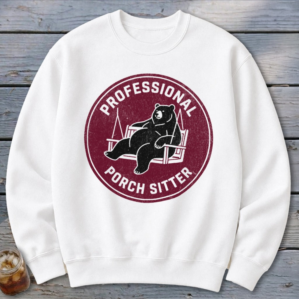 A white crewneck sweatshirt featuring a maroon circular graphic of a bear relaxing on a porch swing, with the text Professional Porch Sitter.
