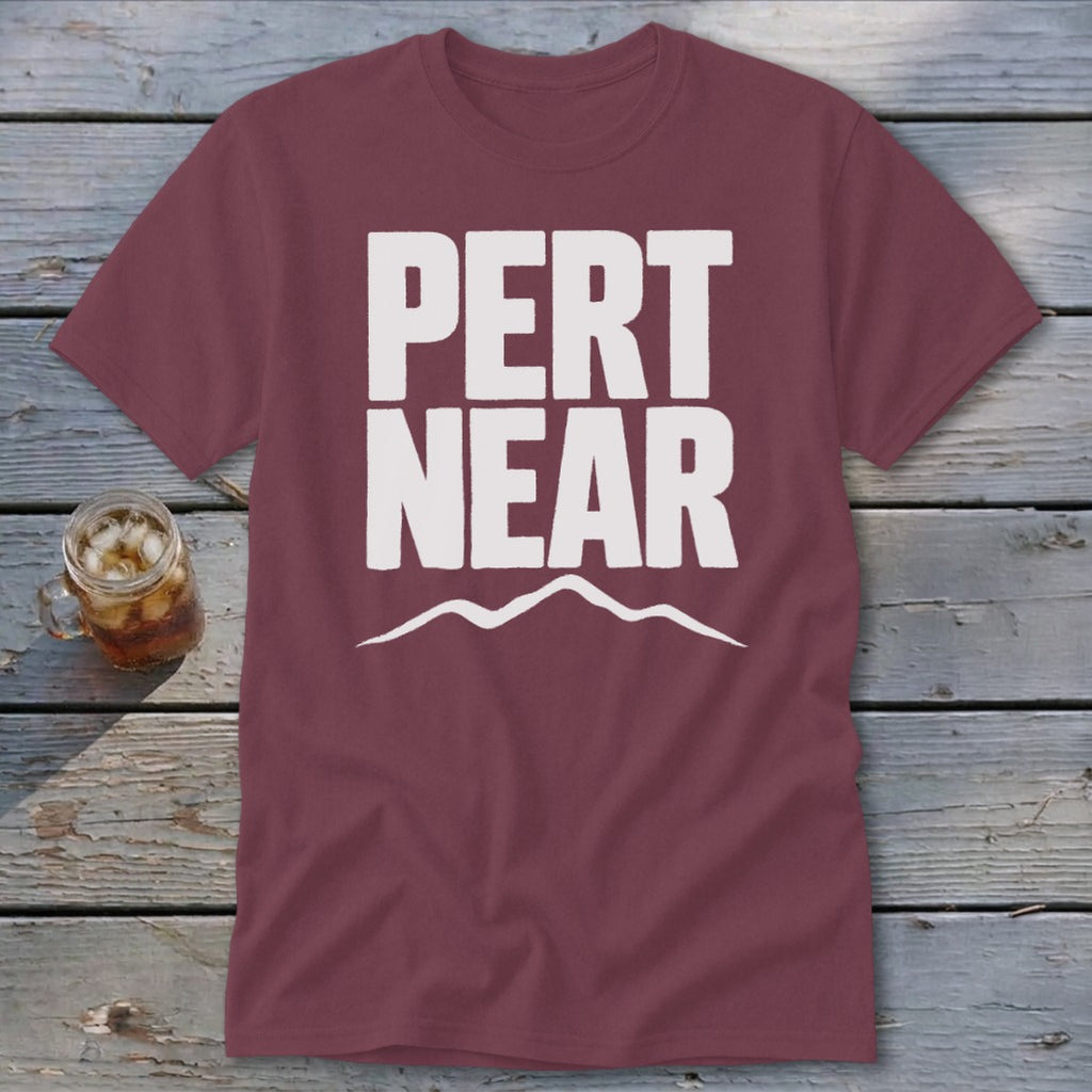 A maroon t-shirt featuring the white text PERT NEAR above a simple mountain outline, resting on weathered wood.