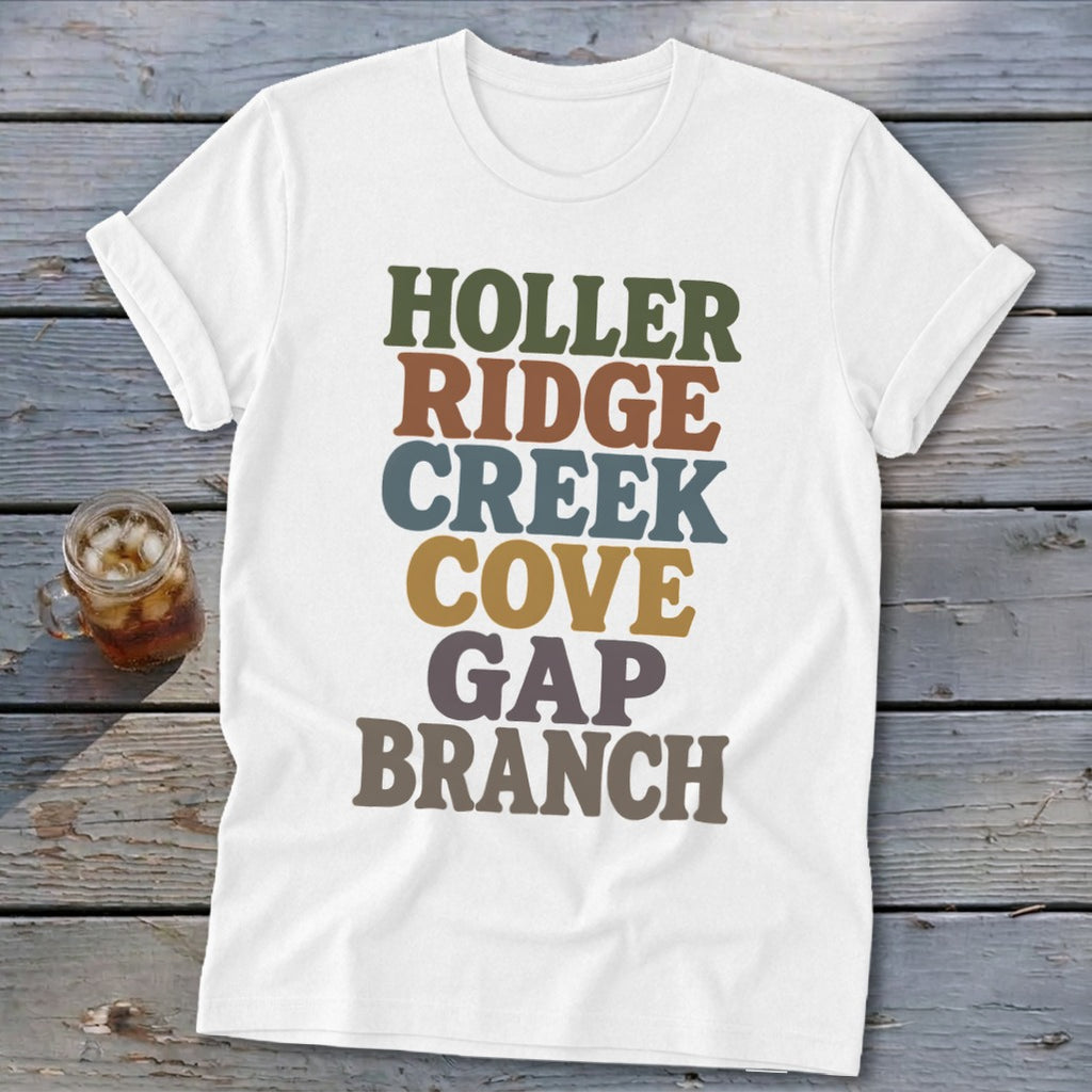 A white t-shirt with rolled sleeves features stacked, colorful retro typography spelling out Appalachian place-name suffixes on a wooden background.