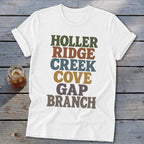 A white t-shirt with rolled sleeves features stacked, colorful retro typography spelling out Appalachian place-name suffixes on a wooden background.