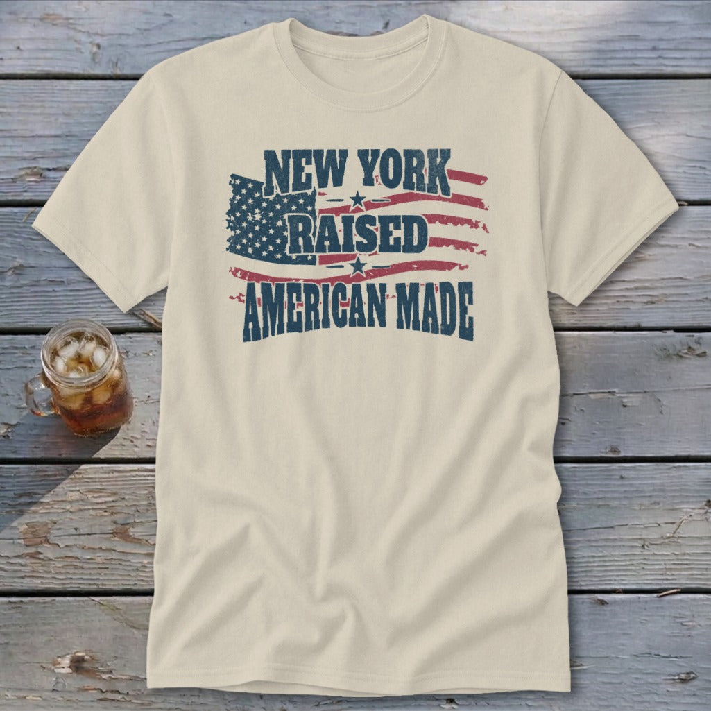 A cream-colored short-sleeve t-shirt featuring a distressed graphic with the text New York Raised American Made over a stylized American flag on a wooden background.