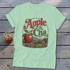 A mint green t-shirt with a vintage graphic featuring an apple, mountains, a cabin, and text.