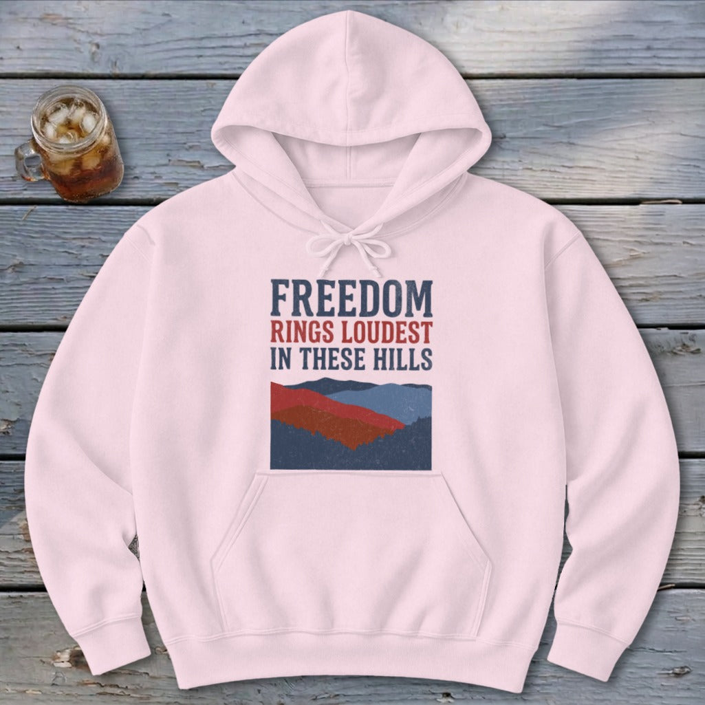 A light pink hooded sweatshirt with a graphic reading FREEDOM RINGS LOUDEST IN THESE HILLS over layered mountain silhouettes.
