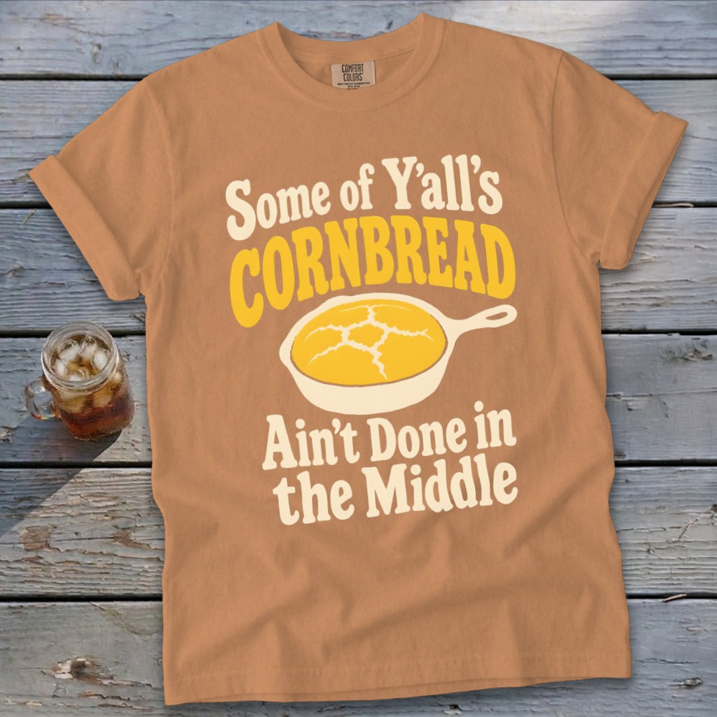 An orange t-shirt featuring text and a skillet graphic that says Some of Y'all's Cornbread Ain't Done in the Middle.