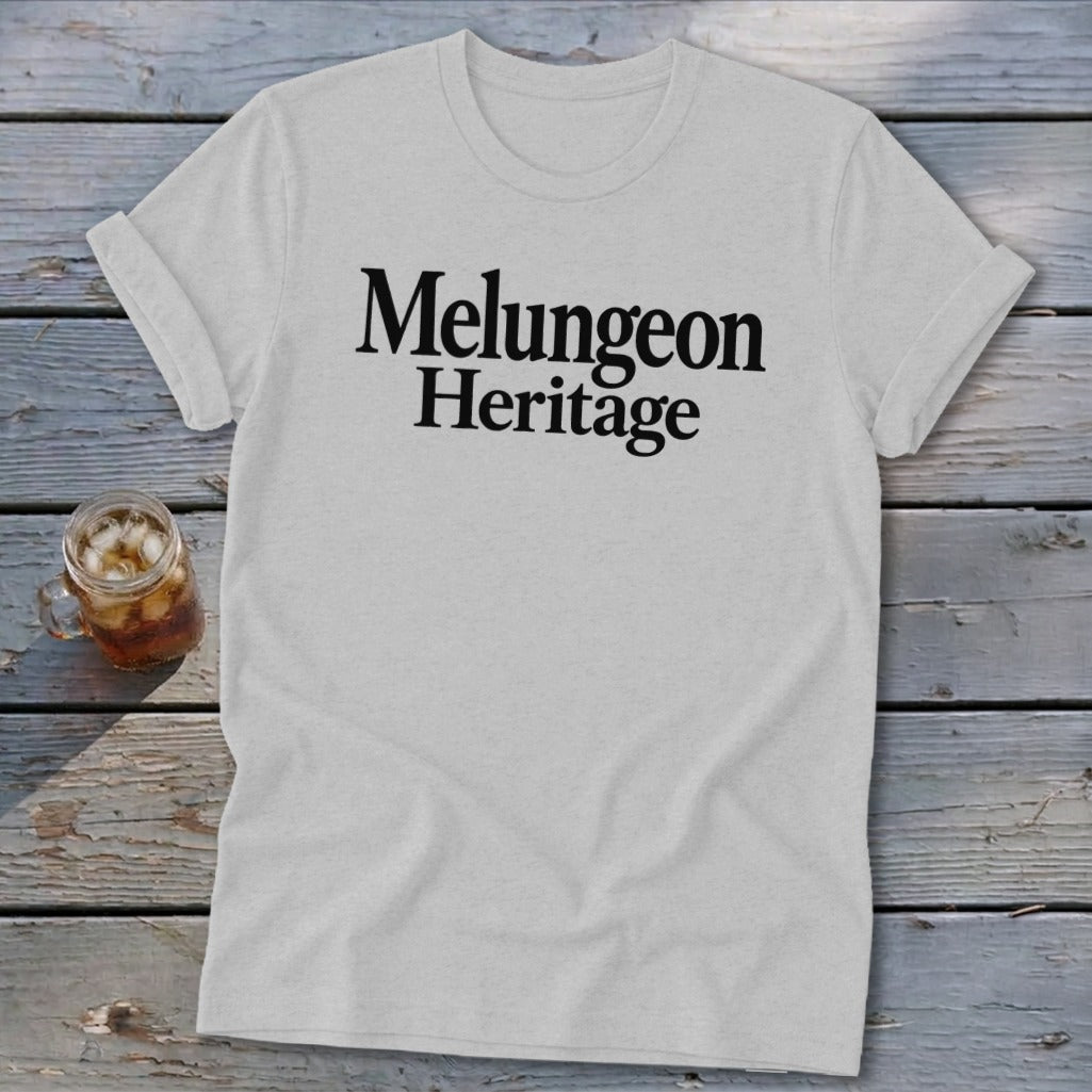 A light gray short-sleeve t-shirt with black text reading Melungeon Heritage lies on a weathered wooden surface, next to a small glass jar filled with iced tea.