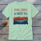 Light green t-shirt with the text "STARS, STRIPES & SWEET TEA" above a layered mountain and firework graphic.