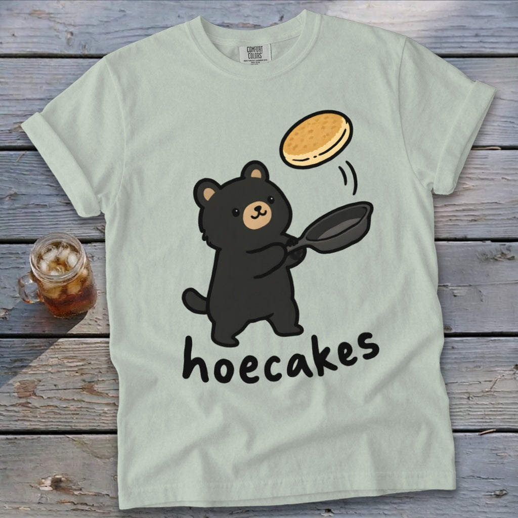 A light green t-shirt features a cartoon black bear flipping a pancake with text below it reading hoecakes.