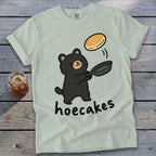 A light green t-shirt features a cartoon black bear flipping a pancake with text below it reading hoecakes.