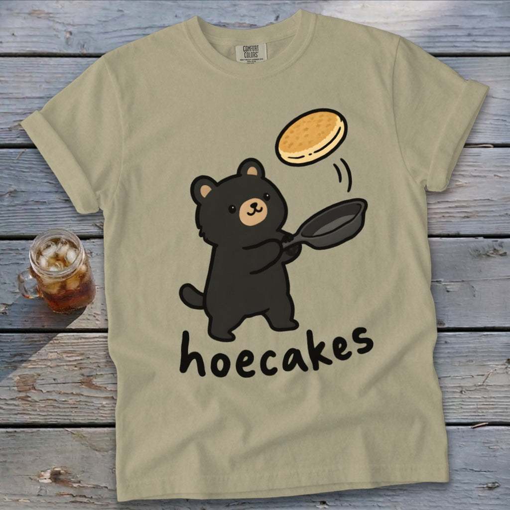 Olive green t-shirt featuring a cute cartoon black bear flipping a pancake with text reading hoecakes.