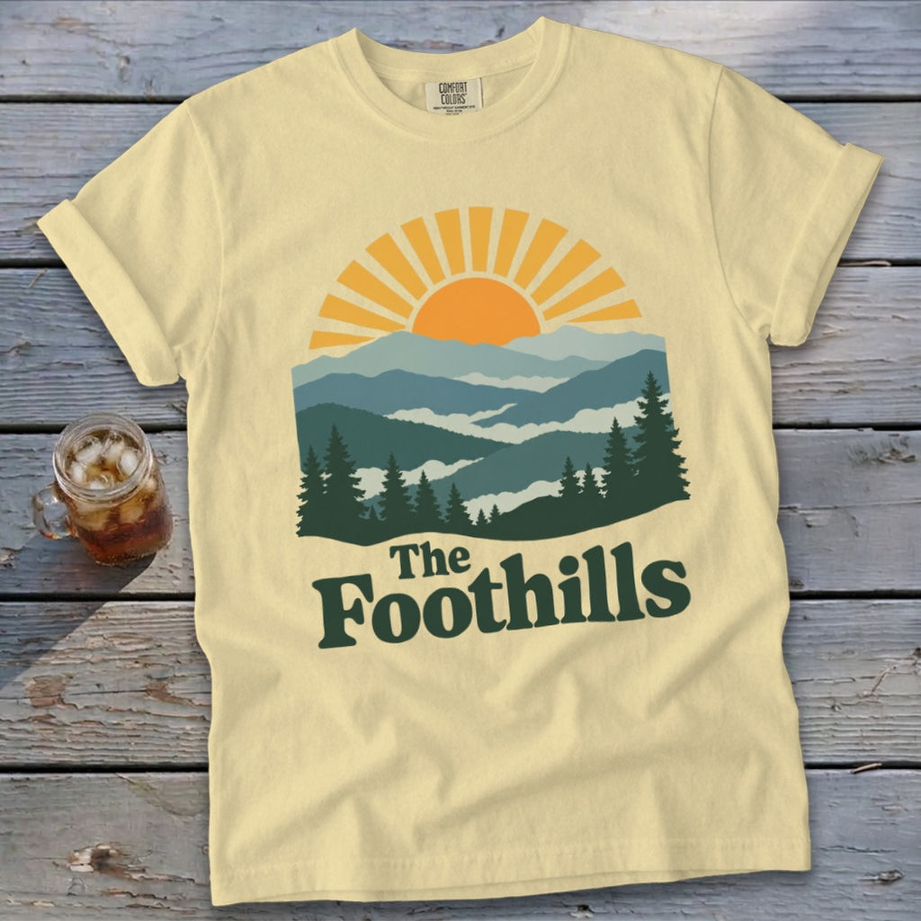 Yellow t-shirt with a graphic of mountains, sun, and trees with "The Foothills" text.