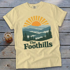 Yellow t-shirt with a graphic of mountains, sun, and trees with "The Foothills" text.