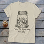 A cream-colored t-shirt with a vintage jar graphic containing a mountain landscape, and the text Carry The Mountains With You.