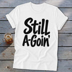 A white t-shirt with rolled sleeves features black text reading "Still A-Goin'".