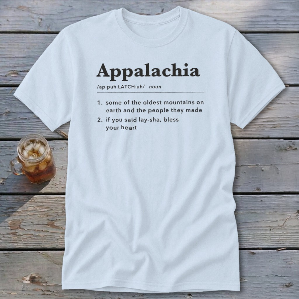 A light blue t-shirt printed with a dictionary definition of Appalachia rests on a wooden surface beside a small jar of iced tea.