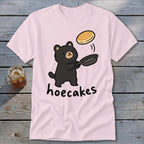 Light pink t-shirt featuring a cartoon black bear flipping a pancake from a skillet, with the text hoecakes underneath.