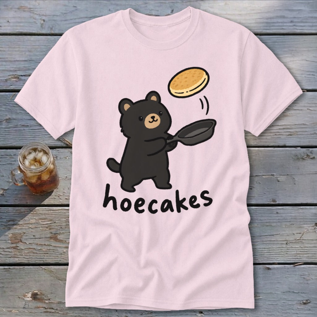 Light pink t-shirt featuring a cartoon black bear flipping a pancake from a skillet, with the text hoecakes underneath.