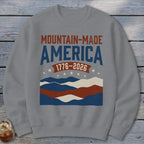 A grey crewneck sweatshirt featuring a distressed graphic with red and blue text, mountain waves, and stars.