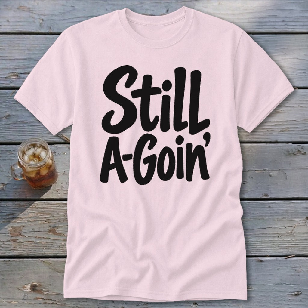 A light pink short-sleeved t-shirt with the black text "Still A-Goin'" lies flat on a weathered wooden surface, accompanied by a glass of iced tea.